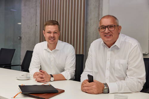 Zwei lächelnde Geschäftsleute in weißen Hemden sitzen am Konferenztisch, einer mit Notizbuch und Kaffeetasse, der andere mit Smartphone in einem modernen Besprechungsraum.