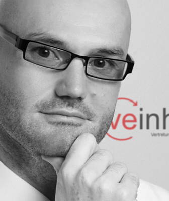 Nachdenklicher glatzköpfiger Mann mit Brille stützt sein Kinn mit der Hand vor einem unscharfen Firmenlogo im Hintergrund