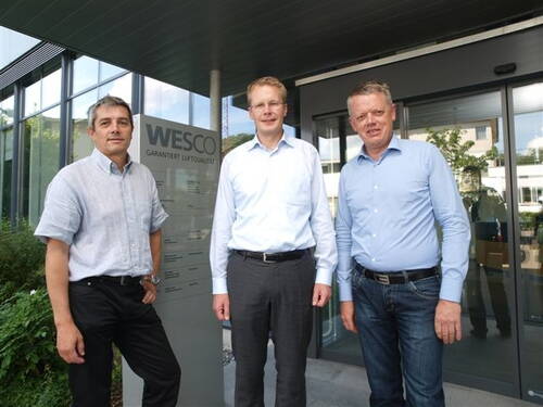 Drei Männer in Business-Kleidung stehen vor dem Eingang eines modernen Firmengebäudes mit WESCO-Firmenschild im Hintergrund