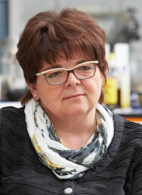 Mittleren Alters Frau mit kurz geschnittenen braunen Haaren, Brille und gemustertem Schal sitzt nachdenklich in einem Innenraum und blickt leicht zur Seite