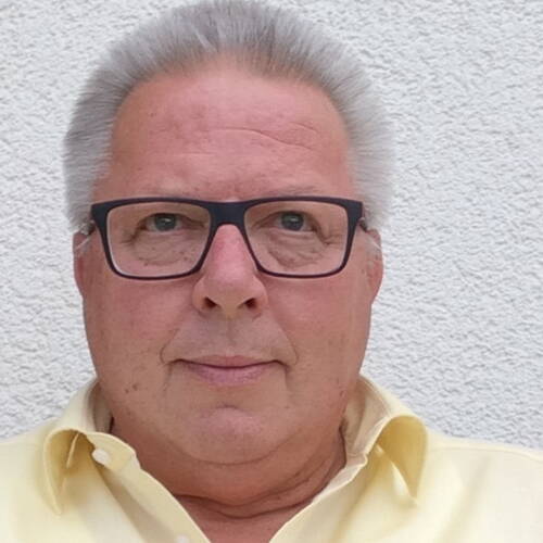 Älterer Mann mit grauem, nach hinten gekämmtem Haar und dunkler Brille blickt ernst in die Kamera, vor einer hellen Putzwand und in ein gelbes Hemd gekleidet.