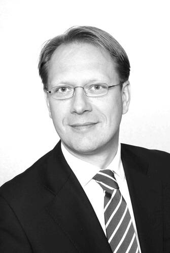 Porträt eines lächelnden Mannes mit Brille im Anzug und gestreifter Krawatte vor hellem Hintergrund