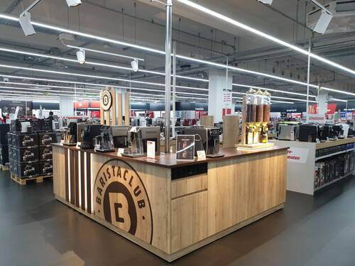 Moderne BaristaClub-Kaffeetheke aus hellem Holz in einem Elektronikmarkt mit Kaffeemaschinen, Bohnen-Spendern und Präsentationsfläche für Kaffeespezialitäten