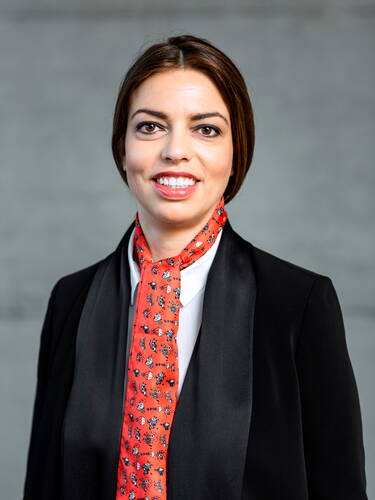 Lächelnde Geschäftsfrau mit dunkelbraunen Haaren im schwarzen Blazer und weißem Hemd, die einen auffälligen roten Schal mit kleinem Muster vor grauem Hintergrund trägt