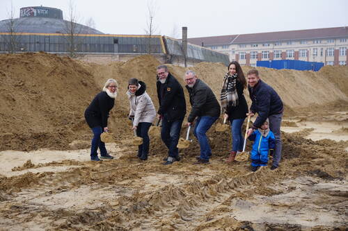 Mehrere Erwachsene und ein kleines Kind stehen mit Spaten in einer sandigen Baugrube und heben lächelnd Erde aus, im Hintergrund sind hohe Sandhügel und industrielle Gebäude zu sehen.