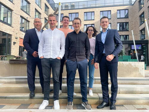 Sechs lächelnde Kolleginnen und Kollegen in Freizeit- und Businesskleidung stehen auf einer Treppe vor einem modernen Bürogebäude mit großen Fenstern und Backsteinfassade.