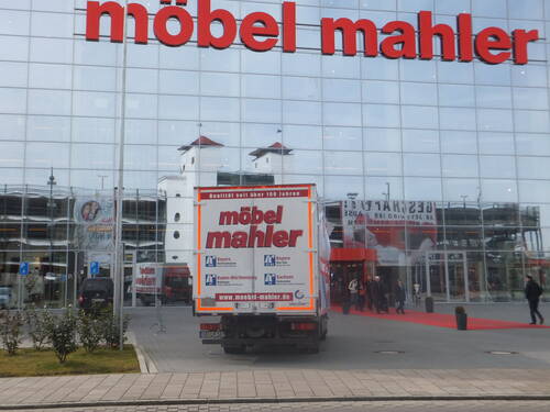 Lieferwagen von „möbel mahler“ fährt auf den Eingang eines großen gläsernen Möbelhauses mit rotem Teppich zu