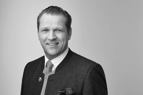 Lächelnder Mann mittleren Alters im traditionellen dunklen Sakko mit Krawatte und Einstecktuch vor hellem, neutralem Hintergrund in einer Schwarzweiß-Porträtaufnahme.
