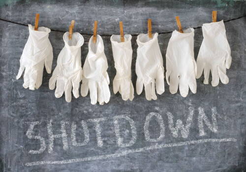 Weiße Einweghandschuhe hängen an einer Wäscheleine vor einer Tafel, auf der groß das Wort „SHUTDOWN“ mit Kreide geschrieben ist.