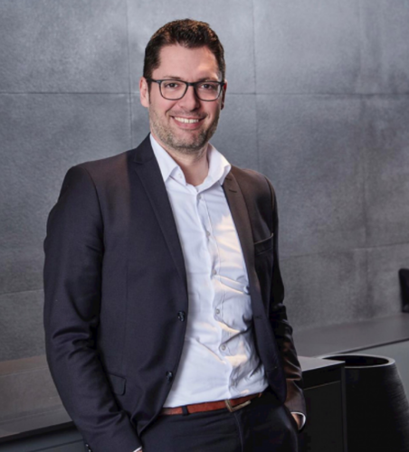 Lächelnder Mann im Business-Anzug mit Brille lehnt sich in modernem Büro an eine dunkle Theke