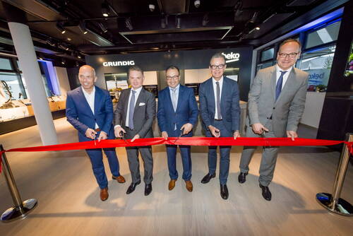 Fünf Geschäftsleute im Anzug schneiden gemeinsam ein rotes Band zur feierlichen Eröffnung eines modernen Showrooms mit Grundig- und Beko-Branding durch