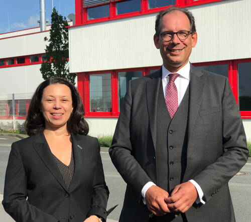 Zwei Geschäftsleute im Anzug lächeln nebeneinander im Sonnenlicht vor einem modernen Industriegebäude mit roten Fensterrahmen.