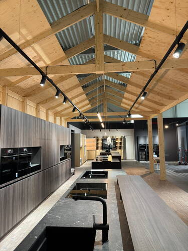 Modernes Küchenstudio mit offener Holztragwerksdecke, langen Kochinseln mit Induktionsfeldern und eingebauten schwarzen Einbaugeräten entlang der Wand