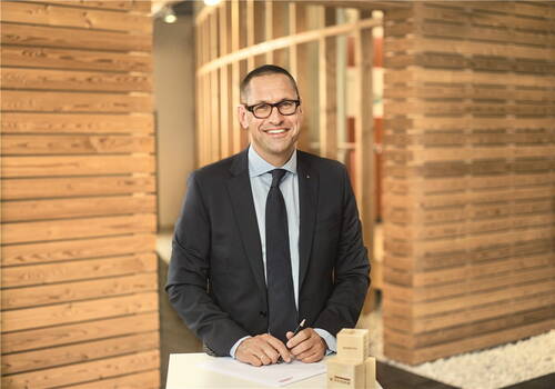 Lächelnder Geschäftsmann im Anzug steht in modernem Holz-Büro und unterschreibt Dokumente an einem weißen Stehpult