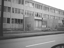 Mehrstöckiger Plattenbau mit heruntergekommenen Fassaden, Bauzäunen und leeren Eingangsbereichen an einer breiten, fast leeren Straße.
