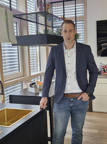 Mann im Sakko und Jeans steht entspannt in einer modernen Küche mit goldenem Spülbecken und großen Fensterfronten