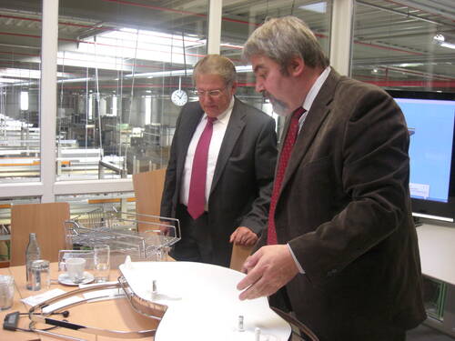 Zwei Männer im Anzug begutachten in einem Besprechungsraum technische Bauteile und Metallkörbe vor einer Glaswand mit Blick in eine industrielle Produktionshalle