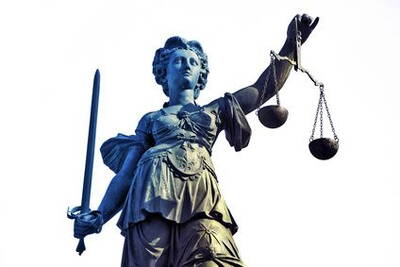 Bronzene Justitia-Statue hält ein Schwert in der rechten Hand und eine Waage in der linken vor hellem Himmel