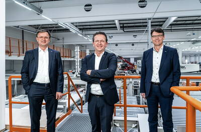 Drei Geschäftsleute in Anzügen stehen lächelnd auf einer erhöhten Plattform in einer modernen Fabrikhalle mit Maschinen im Hintergrund