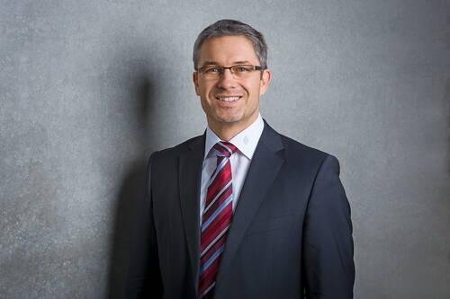 Geschäftsmann mit Brille und Anzug lächelt vor grauer Betonwand in professionellem Porträt.