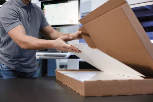 Mitarbeiter in grauem Poloshirt legt einen großen, flachen Druck sorgfältig in einen geöffneten Versandkarton vor einem Computerarbeitsplatz.