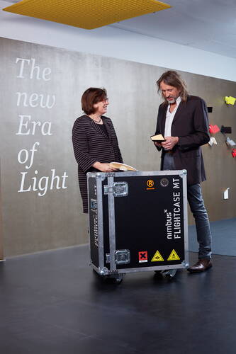 Zwei Personen im modernen Showroom unterhalten sich über Musterteile an einem großen schwarzen Flightcase mit der Aufschrift „Nimbus Flightcase MT“, neben einer Wand mit dem Slogan „The new Era of Light“ und farbigen Leuchten.