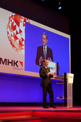 Redner im Anzug spricht an einem transparenten Pult auf einer Bühne, während sein vergrößertes Abbild hinter ihm auf einer Leinwand mit MHK-Group-Logo projiziert wird