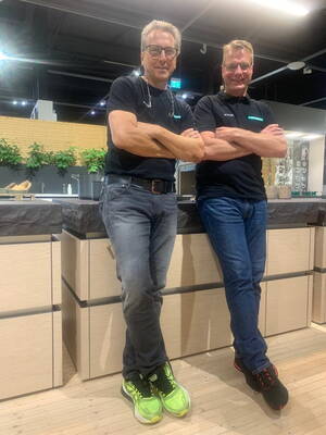 Zwei lässig an einer modernen Kücheninsel angelehnte Männer in Arbeitskleidung, die mit verschränkten Armen und auffälligen Sportschuhen in einem hell beleuchteten Showroom posieren