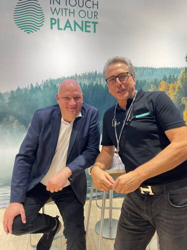 Zwei lächelnde Männer posieren entspannt an einem Messestand vor einem Waldmotiv und dem Schriftzug „In Touch With Our Planet“.