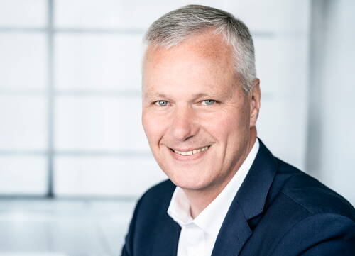 Älterer Mann mit grauen Haaren im dunklen Anzug lächelt freundlich in die Kamera vor einem hellen, unscharfen Bürohintergrund.