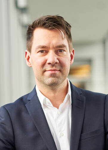 Mann im dunkelblauen Anzug mit weißem Hemd blickt freundlich und direkt in die Kamera vor einem modernen, hellen Bürohintergrund.