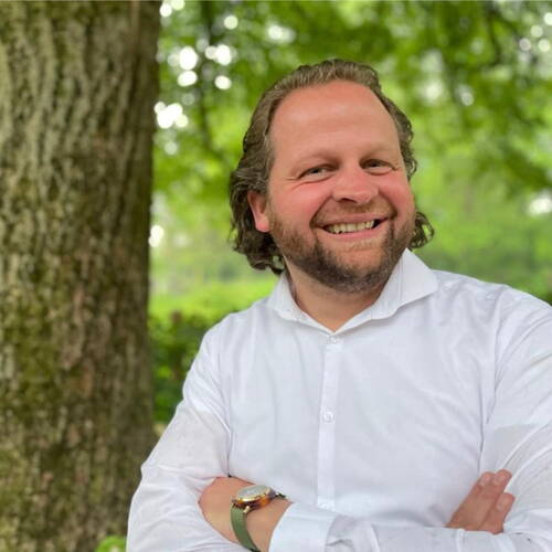 Lächelnder Mann mit Bart und weißem Hemd steht mit verschränkten Armen vor einem Baum in einem grünen Park.