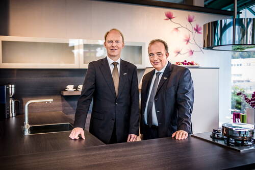 Zwei Geschäftsleute im Anzug stehen lächelnd an einer modernen Kücheninsel mit Kochfeld und Spüle in einem hellen Showroom.