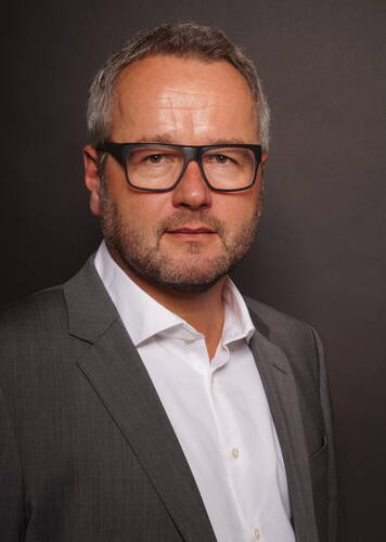 Mittelalterlicher Mann mit grauem Haar, dunkler Brille und Dreitagebart trägt grauen Anzug und weißes Hemd vor dunkelgrauem Hintergrund und blickt ernst in die Kamera.