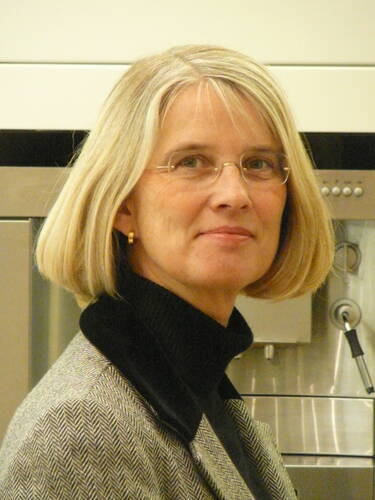 Ältere Frau mit blondem Bob und Brille im Profil, die in eleganter Jacke und Rollkragenpullover vor einer Edelstahlküche freundlich in die Kamera blickt.