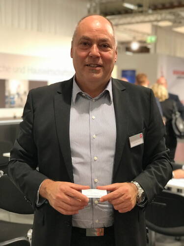 Geschäftsmann im Anzug hält seine Visitenkarte in den Händen und lächelt in einer modernen Messe- oder Konferenzumgebung in die Kamera.