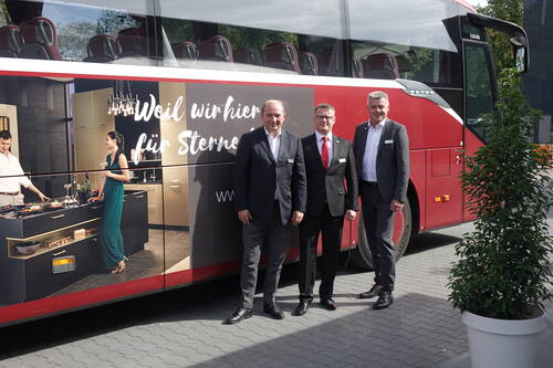 Drei Geschäftsleute im Anzug posieren vor einem roten Reisebus mit großflächiger Küchenwerbung und dem Slogan „Weil wir hier für Sterne …“ auf der Fahrzeugseite