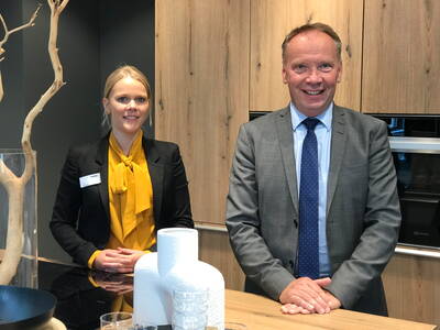 Zwei lächelnde Geschäftsleute stehen nebeneinander in einer modernen Küche, die Frau im schwarzen Blazer mit gelber Bluse, der Mann im grauen Anzug mit blauer Krawatte, vor hellen Holzfronten und Küchenaccessoires auf der Arbeitsplatte.