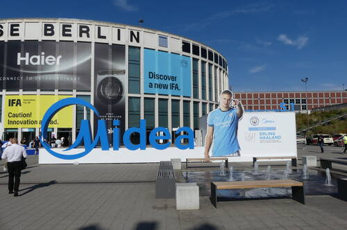 Große Midea-Werbeinstallation vor dem Messegelände Berlin mit Haier- und IFA-Bannern sowie lebensgroßer Fußballerfigur neben Springbrunnen und Besuchern im Eingangsbereich