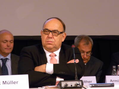 Älterer Mann im Anzug mit verschränkten Armen sitzt ernst vor einem Mikrofon in einer Podiumsdiskussion, während zwei weitere Männer im Hintergrund in Unterlagen schauen.