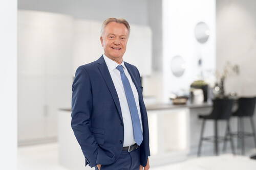 Älterer Geschäftsmann im blauen Anzug steht lächelnd in einem modernen, hellen Büro- oder Küchenraum mit unscharfem Hintergrund.