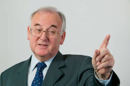 Älterer Mann im Anzug und mit Brille hebt mahnend den Zeigefinger und spricht, als würde er einen wichtigen Punkt betonen.
