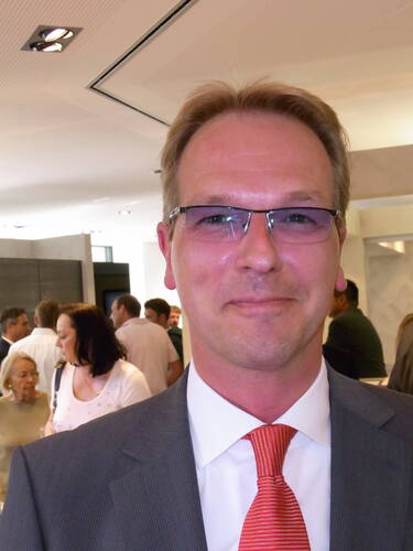 Mittelalterlicher Mann im grauen Anzug mit roter Krawatte lächelt in die Kamera, während im Hintergrund eine Gruppe von Menschen in einem hellen Veranstaltungsraum miteinander spricht