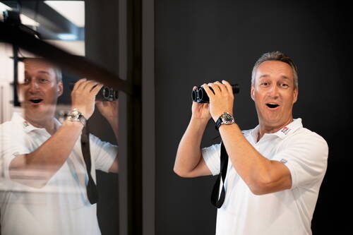Mittelalter Mann im weißen Poloshirt hält ein Fernglas vor sich, schaut überrascht hinein und spiegelt sich lächelnd in einer Glaswand.