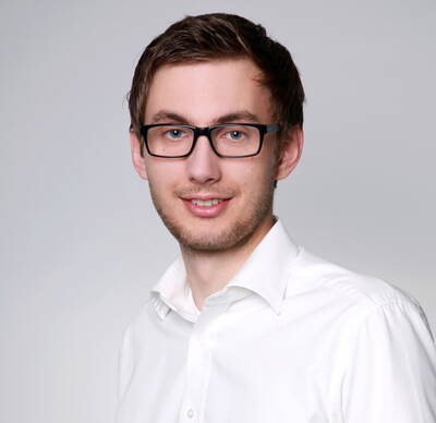 Junger Mann mit dunkler Brille und weißem Hemd lächelt vor neutral grauem Hintergrund in die Kamera