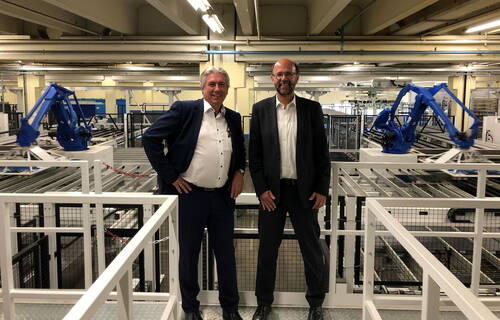 Zwei Männer in Anzügen stehen lächelnd auf einer Plattform in einer modernen Fabrikhalle mit blauen Industrierobotern und Förderanlagen im Hintergrund