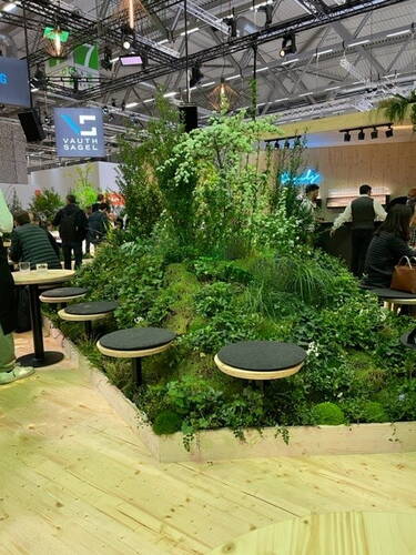 Runde Sitzhocker umgeben eine üppig bepflanzte, hügelartige Grüninsel in einer modernen Messehalle mit Besuchern im Hintergrund