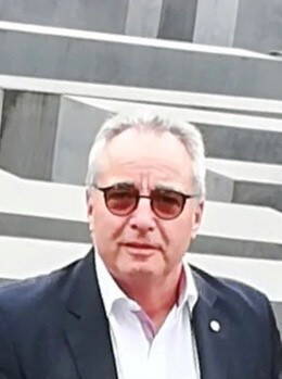 Älterer Mann mit grauen Haaren, Sonnenbrille und dunklem Sakko steht vor grauen, stufenartig angeordneten Betonblöcken.