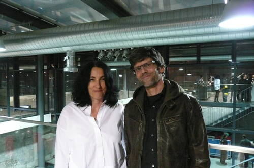 Zwei Menschen stehen lächelnd nebeneinander in einem modernen Industriegebäude mit Glasgeländern und sichtbaren Lüftungsrohren, während im Hintergrund eine Gruppe Personen bei einer Veranstaltung zusammensteht.