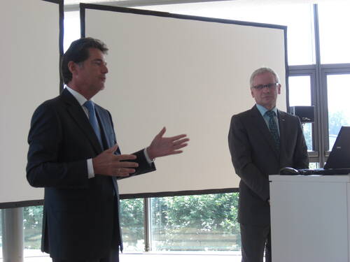 Zwei Geschäftsleute im Anzug halten eine Präsentation in einem hellen Konferenzraum, während einer spricht und gestikuliert und der andere neben einem Laptop am Rednerpult steht.